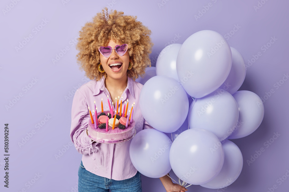 Positive curly haired woman holds festive cake with burning candles bunch of inflated balloons laughs out gladfully wears heart shaped sunglasses shirt and jeans poses indoor. Special occasion Positive curly haired woman holds festive cake with burning candles bunch of inflated balloons laughs out gladfully wears heart shaped sunglasses shirt and jeans poses indoor. Special occasion