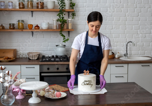 Woman in an apron holds white cake with the inscription Happy birthday and the numbers 32 in her hands and puts it on cardboard box. Packaging for delivery. Selective focus.