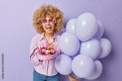 Positive curly haired woman holds festive cake with burning candles bunch of inflated balloons laughs out gladfully wears heart shaped sunglasses shirt and jeans poses indoor. Special occasion