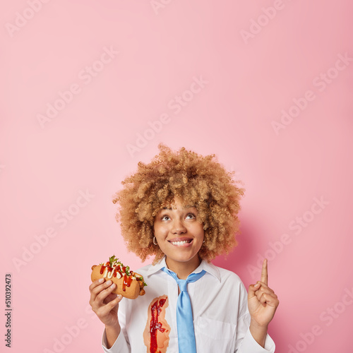 Fotografia Cheerful woman with curly bushy hair eats delicious hot dog harmful food points