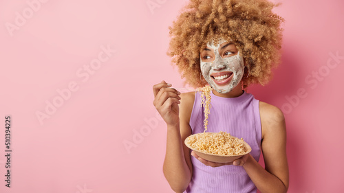 Horizontal shot of glad young woman eats pasta from bowl looks happily away undergoes beauty procedures applies clay nourishing mask on face isolated on pink background empty space for your text