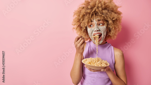 Indoor shot of beautiful curly haired woman applies beauty mask for reducing blackheads and fine lines eats appetizing pasta looks away stands against pink studio wall empty space for your promo