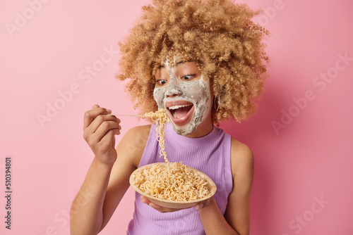 Indoor shot of cheerful curly haired woman eats noodle feels hungry dressed in casual purple t shirt applies nourishing clay mask to reduce fine lines isolated over pink background. Beauty procedures