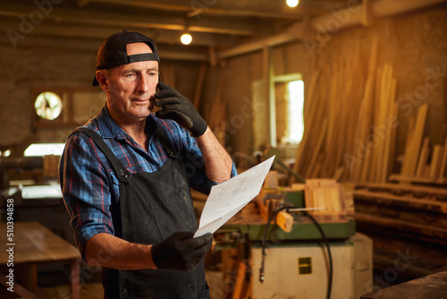 Portrait of a senior carpenter looking at blueprints plans and talking phone with a client in the carpentry workshop
