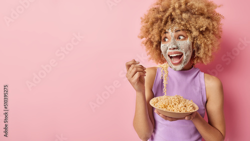 Excited cheerful young woman with curly hair applies beauty mask undergoes cosmetic procedurs looks amazed away dressed casually eats spaghetti for lunch isolated over pink wall blank space.