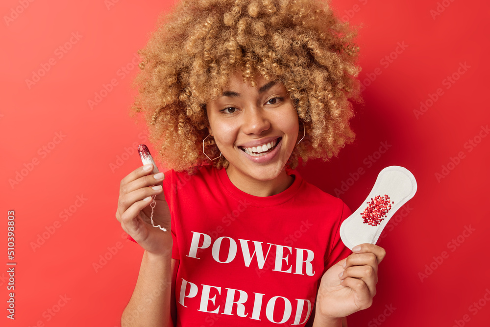 Positive woman with curly hair holds sanitary napkin and tampon chooses product for monthly menstrual cycle cares about protection and women health wears casual t shirt isolated over red background Positive woman with curly hair holds sanitary napkin and tampon chooses product for monthly menstrual cycle cares about protection and women health wears casual t shirt isolated over red background