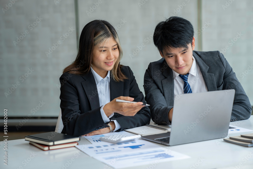 Two beautiful Asian businessmen working together in the office sitting at desk with a laptop working on paperwork to discuss the project's financial reports. business organization cooperation concept Two beautiful Asian businessmen working together in the office sitting at desk with a laptop working on paperwork to discuss the project's financial reports. business organization cooperation concept