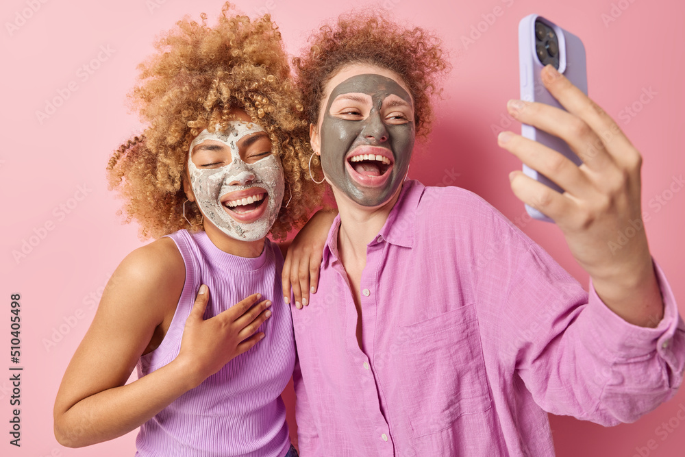 Overjoyed two young women apply beauty facial masks laugh joyfully take selfie via modern smartphone express positive emotions dressed casually isolated over pink background. Skin care concept