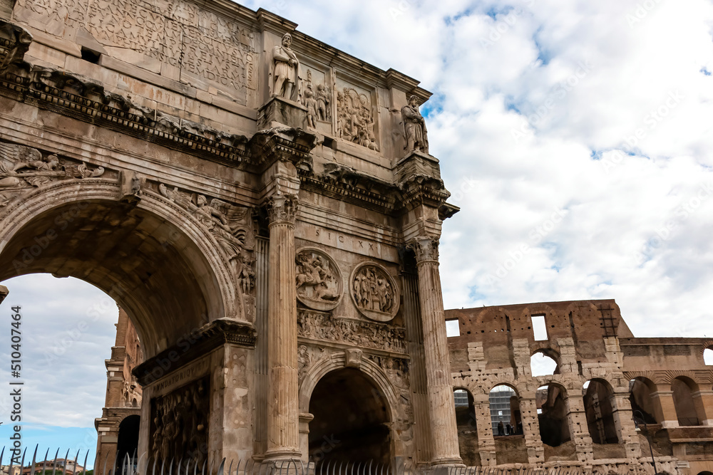 Panoramic view on the exterior of Colosseum and the Triumphal Arch of Constantine in city of Rome, Lazio, Italy, Europe. UNESCO World Heritage Site. Tourism in European city. Clouds over the landmark