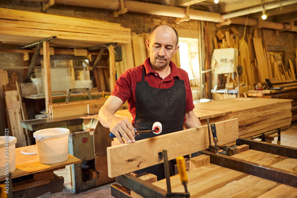 Portrait of a senior carpenter in uniform gluing wooden bars with hand pressures at the carpentry manufacturing Portrait of a senior carpenter in uniform gluing wooden bars with hand pressures at the carpentry manufacturing