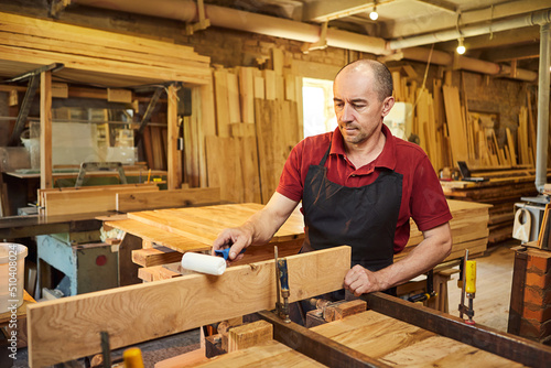Portrait of a senior carpenter in uniform gluing wooden bars with hand pressures at the carpentry manufacturing