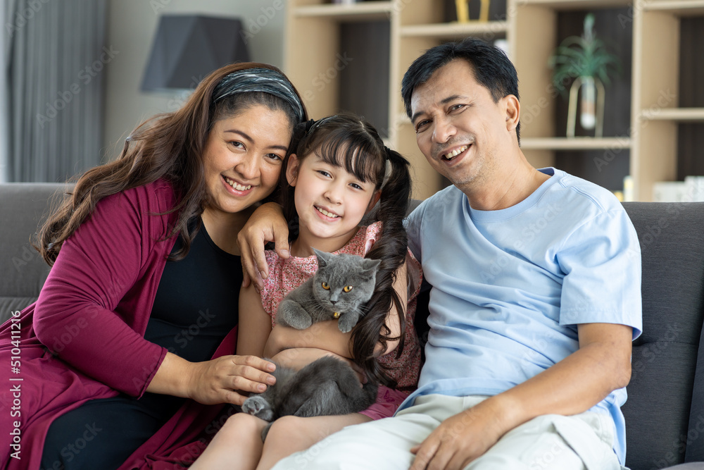 Portrait of diverse asian family couple with child daughter playing with pet cat on sofa in living room at home. Smiling parents and teen girl kid embracing cute cat while sitting on sofa.