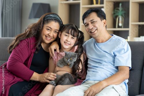 Portrait of diverse asian family couple with child daughter playing with pet cat on sofa in living room at home. Smiling parents and teen girl kid embracing cute cat while sitting on sofa.