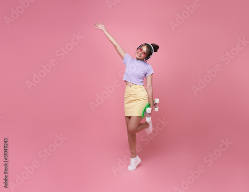 Young asian teenage girl holding skateboard with wearing wireless headphones listening to music on pink background.