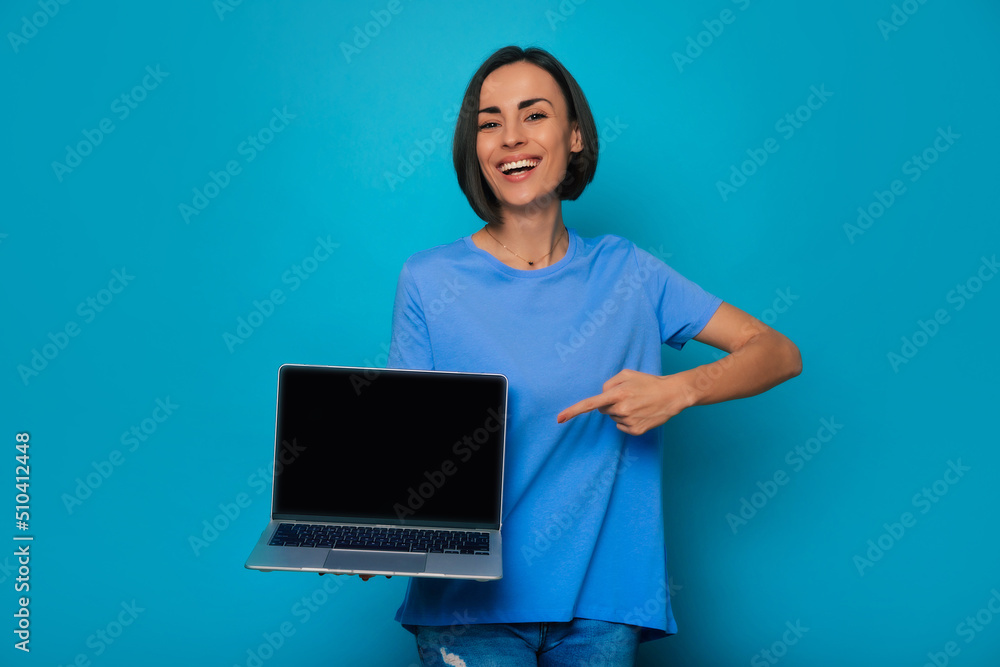 Attractive smiling brunette woman is presenting the modern laptop with blank screen and pointing on it while she isolated on blue background. New device for work and business Attractive smiling brunette woman is presenting the modern laptop with blank screen and pointing on it while she isolated on blue background. New device for work and business