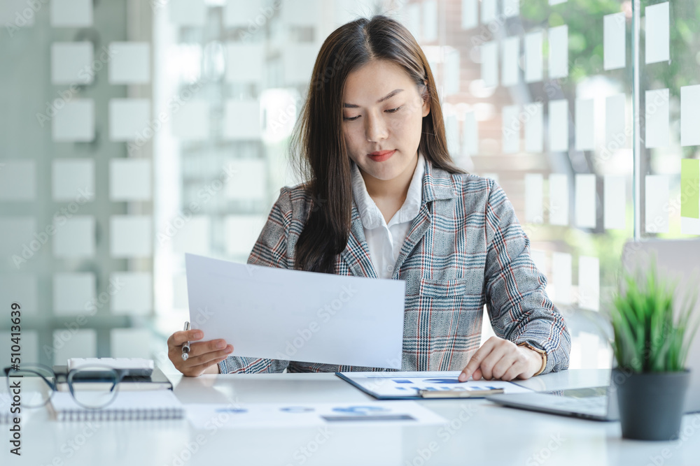 Smiling asian business woman sitting at office desk with computer, holding financial documents, paper reports, satisfied with new project first results. Positive hr manager looking through resume. Smiling asian business woman sitting at office desk with computer, holding financial documents, paper reports, satisfied with new project first results. Positive hr manager looking through resume.