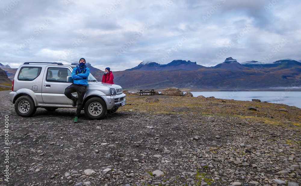 A couple standing on the sides of a car and enjoying the landscape. Road tripping around Iceland. The car is pulled on the small parking lot, next to a fjord shore. Sky is covered with thick clouds.