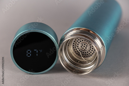 Thermos with open lid and teapot on gray background. Cylindrical lid with LED digital display. Temperature reading 87 degrees Fahrenheit. Vacuum flask of turquoise color.
