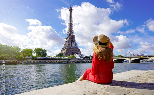 Young traveler woman in red dress and hat sitting on the quay of Seine River looking at Eiffel Tower, famous landmark and travel destination in Paris. © zigres