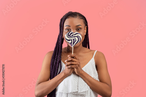 Lovely young black woman covering her mouth with striped heart shaped lollipop on pink studio background