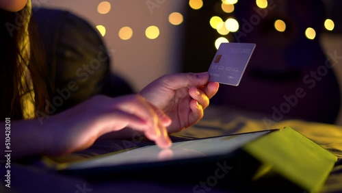 Close up shot of female hand holding credit card using tablet for shopping online lying on bed in bedroom at night.