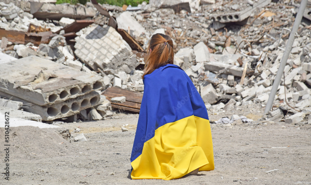 A young girl is on her knees and covered with the national flag of Ukraine against the backdrop of a bombed-out school.