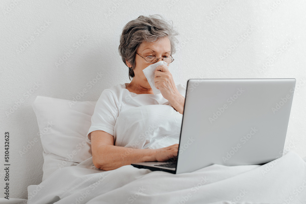 Sick senior woman using laptop while sitting in bed alone in apartment. Elderly woman with runny nose browsing internet or working from home online Sick senior woman using laptop while sitting in bed alone in apartment. Elderly woman with runny nose browsing internet or working from home online