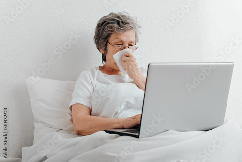 Sick senior woman using laptop while sitting in bed alone in apartment. Elderly woman with runny nose browsing internet or working from home online
