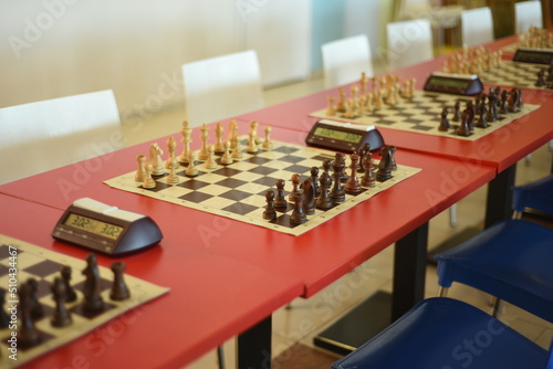 Red tables with chess and chess clocks, white and blue chairs are ready for the tournament.