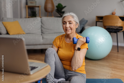 Domestic online training. Senior woman doing exercises with dumbbells in front of laptop, following video tutorial