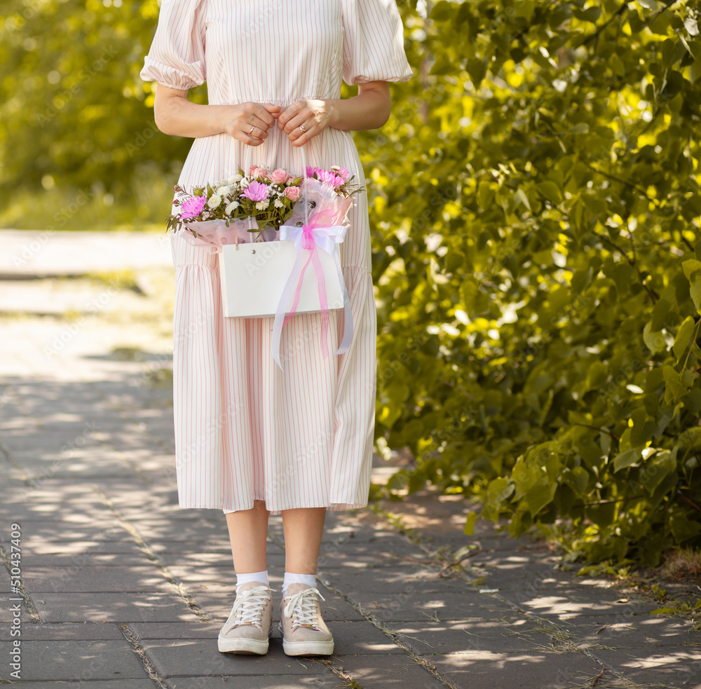 A beautiful bouquet of flowers in a box in the hands of a beautiful girl who walks along the street on a sunny day. Girl in a dress, glasses and sneakers. Focus on the background of flowers. A beautiful bouquet of flowers in a box in the hands of a beautiful girl who walks along the street on a sunny day. Girl in a dress, glasses and sneakers. Focus on the background of flowers.