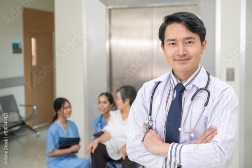 Confident young Asian doctor does crossing hist arm with blurred nurse take care elderly patient in background in hospital.