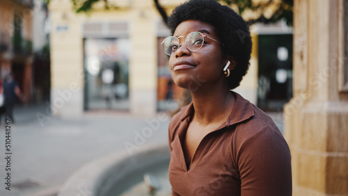 Cute Afro American woman in glasses sitting on the street listen