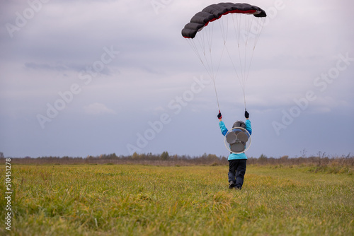 A cute kid in a blue jacket with a propeller and a helmet runs across the green field, trains with a parachute on the wing, holds the brakes, catches the wind, drives a kite and enjoys flying.