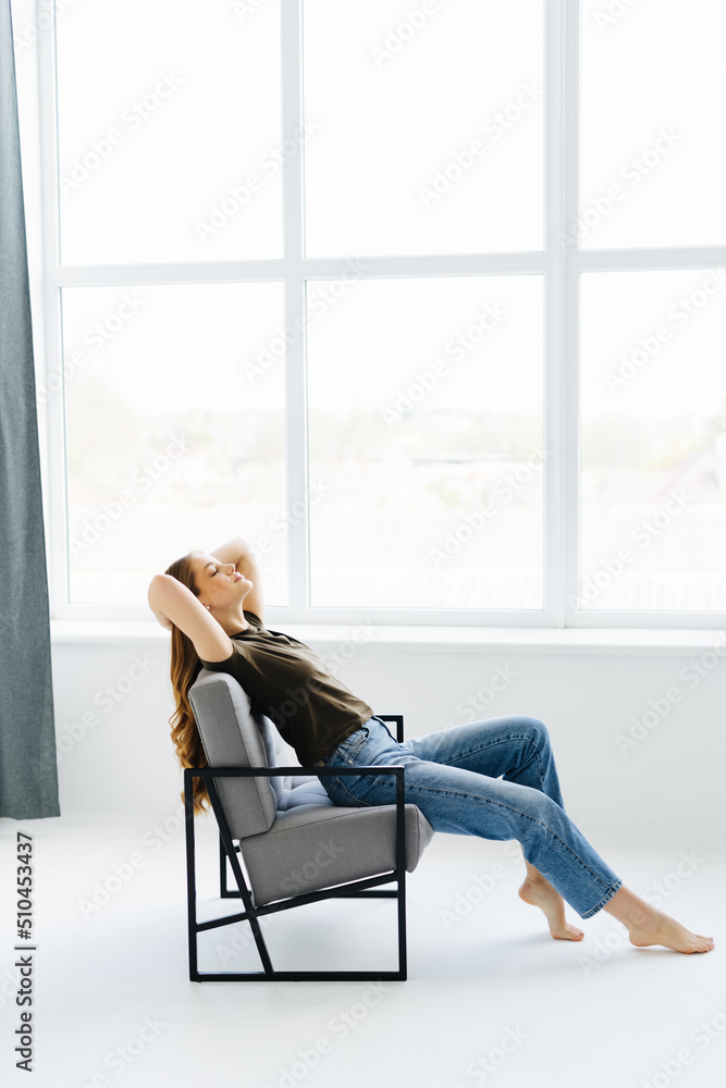 Weekends finally. Relaxed woman is sitting on modern chair near the window in light cozy room at home. She is enjoying peace and morning Weekends finally. Relaxed woman is sitting on modern chair near the window in light cozy room at home. She is enjoying peace and morning