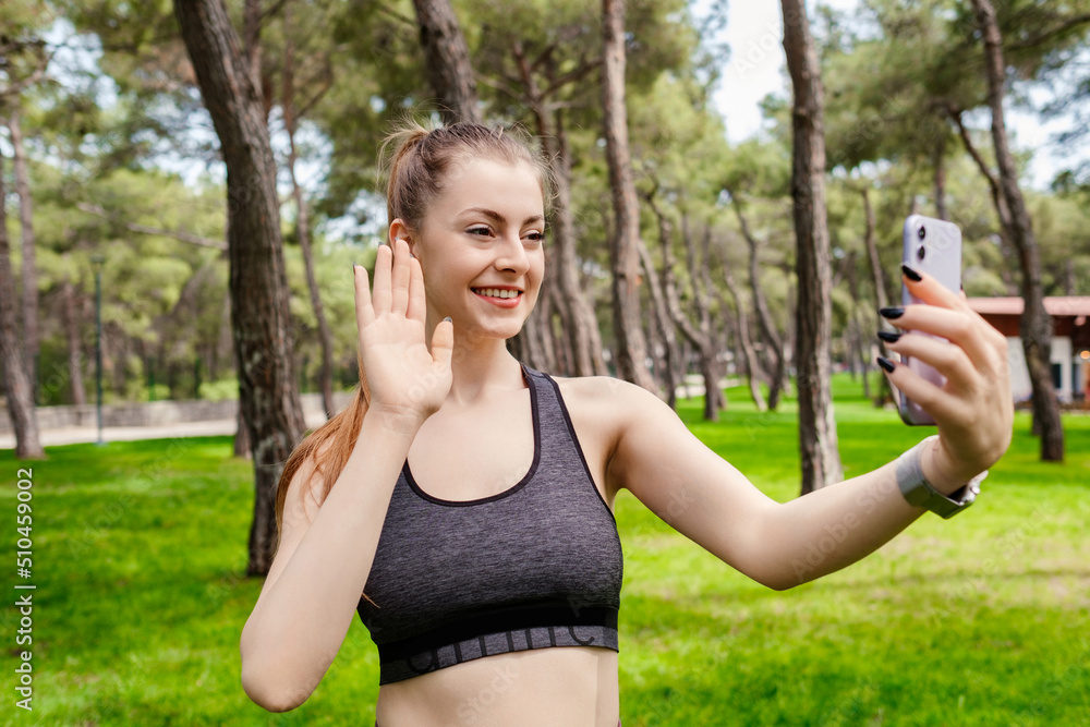Portrait of young brunette woman wearing sportive clothes on city park, outdoors holding phone looking at screen waving hand video calling distance friend online in mobile app using smartphone. Portrait of young brunette woman wearing sportive clothes on city park, outdoors holding phone looking at screen waving hand video calling distance friend online in mobile app using smartphone.