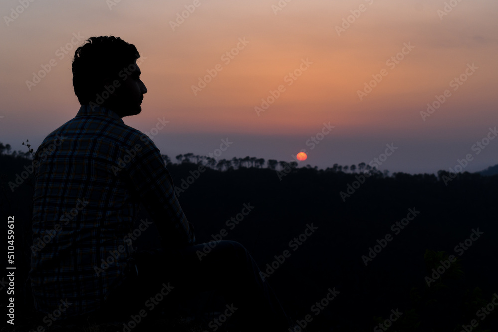 Fototapeta premium Guy sitting looking sideways silhouette pose and sun setting behind mountain chain in background