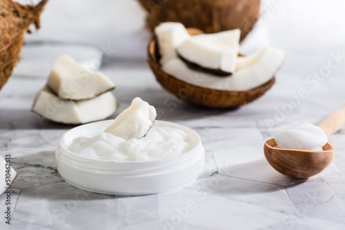 A piece of coconut in coconut oil in a jar and a wooden spoon with oil and coconut on the table.
