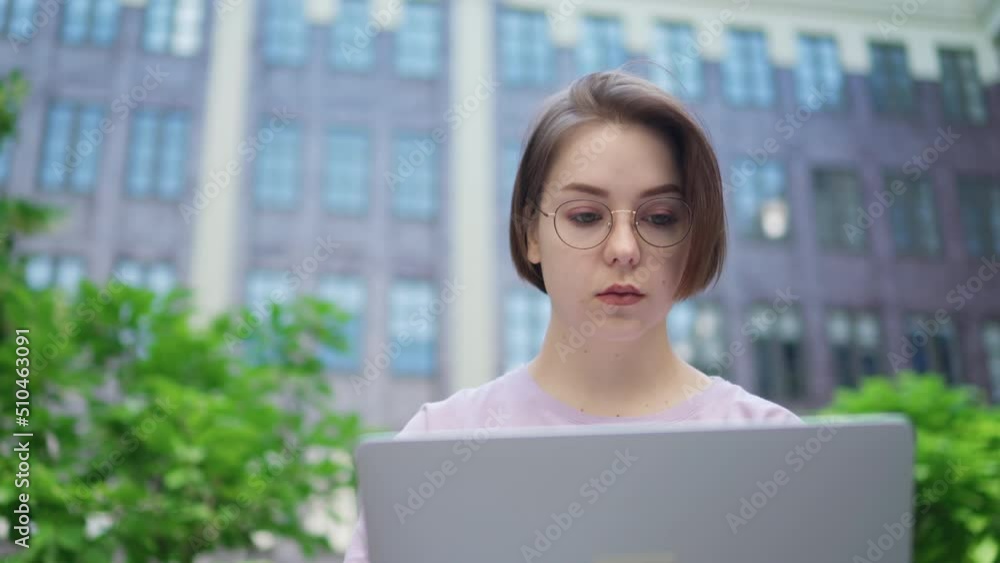 Young Girl with Glasses Looking Into Laptop Screen. Pretty Girl Uses Laptop For Remote Work While Sitting On Bench In Courtyard Of Modern Office Building Young Girl with Glasses Looking Into Laptop Screen. Pretty Girl Uses Laptop For Remote Work While Sitting On Bench In Courtyard Of Modern Office Building