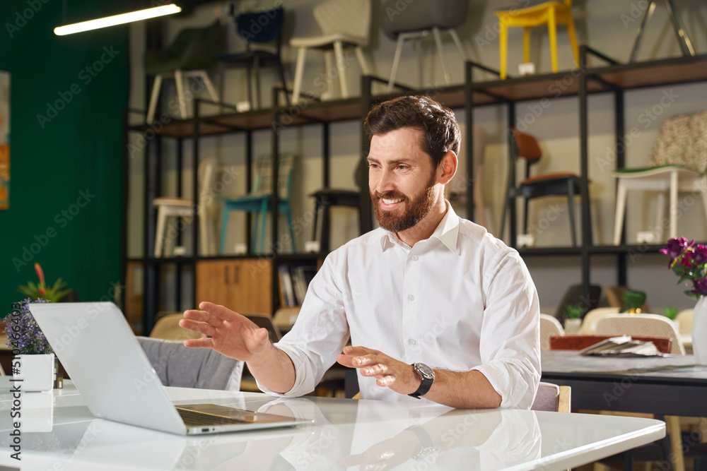 Front view of happy designer, architect sitting at table, working, using laptop. Brunette male with beard looking at screen, smiling, designing. Concept of urban lifestyle. Front view of happy designer, architect sitting at table, working, using laptop. Brunette male with beard looking at screen, smiling, designing. Concept of urban lifestyle.