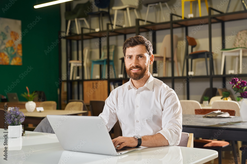 Front view of handsome designer sitting at table, using laptop. Brunette man with beard wearing white shirt typing, looking at camera, smiling. Concept of design and shopping.