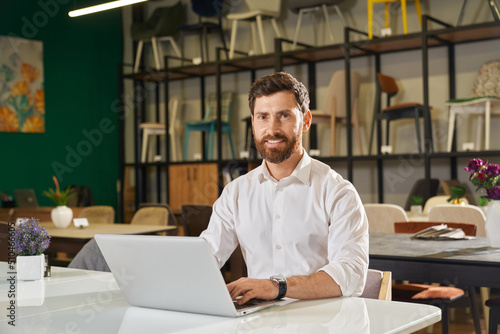 Front view of handsome designer sitting at table, using laptop. Brunette man with beard wearing white shirt typing, looking at camera, smiling. Concept of design and shopping.