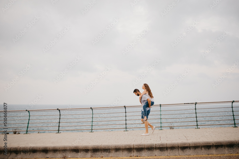 Fototapeta premium A full-length photo of a Hispanic man is carrying on the back his Latina girlfriend through the bridge in a highland park in Spain. A couple of tourists with the sea in the background in Valencia