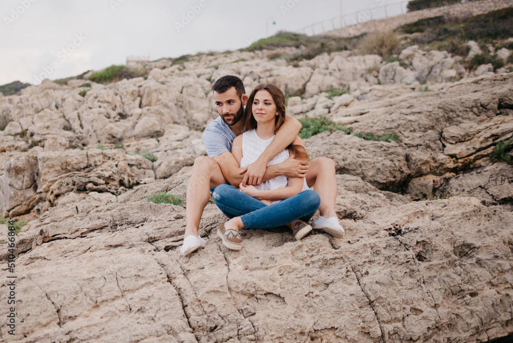 A Hispanic man is holding between his legs his brunette Latina girlfriend while both are sitting on the rocks in Spain. A man is hugging his wife on a date on the coast at sunset in Valencia.