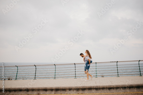 A full-length photo of a Hispanic man is carrying on the back his Latina girlfriend through the bridge in a highland park in Spain. A couple of tourists with the sea in the background in Valencia © Roman Tyukin