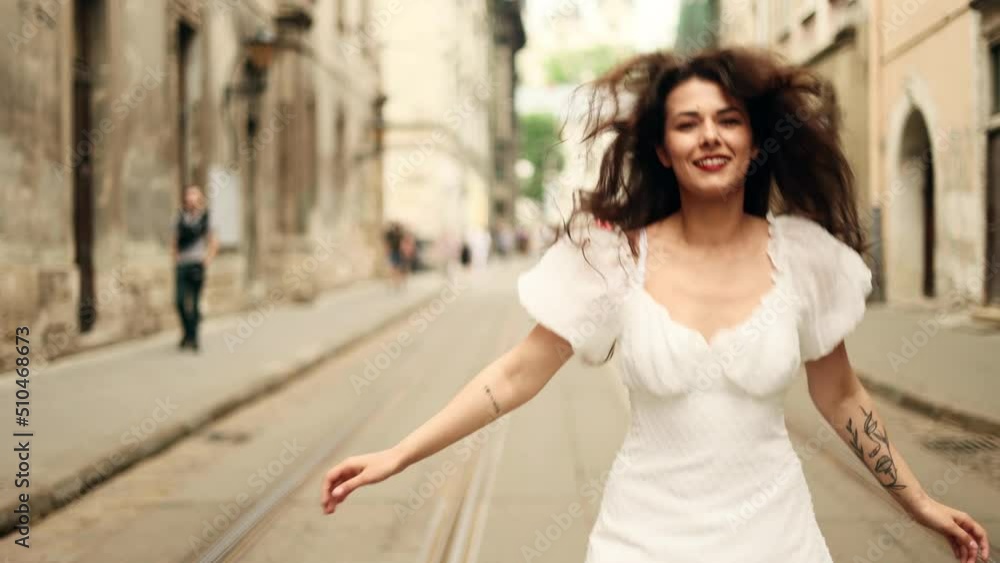 Smiling woman in white dress walking down the city street. Flirty girl with long dark hair turning around in city centre. Smiling woman in white dress walking down the city street. Flirty girl with long dark hair turning around in city centre.