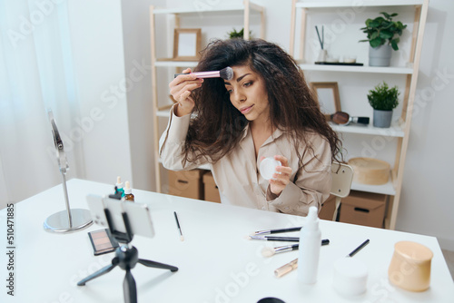 Enthusiastic tanned handsome curly Latin beauty blogger in linen shirt teaches how to use blush in home white interior. Copy space Mockup Banner. Influencer record video review blog using smartphone