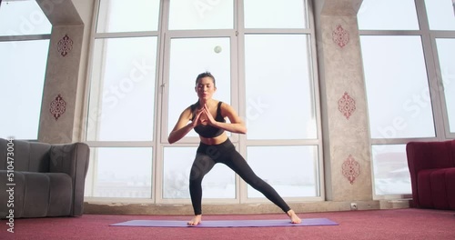 Front view of beautiful girl with ponytail standing alone on yoga mat, squatting, making lunges. Pretty, brunette female wearing sportsuit, doing yoga. Concept of modern life. photo