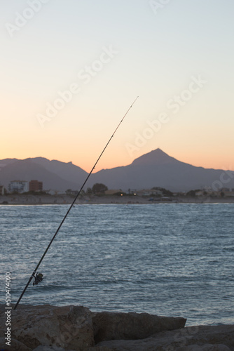 Fishing rod sticks out of jetty rocks at sunset with beach and mountain in the background © Mark