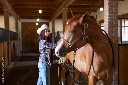 Young girl in cowboy hat combs the stabled horse that looks back at her indoors